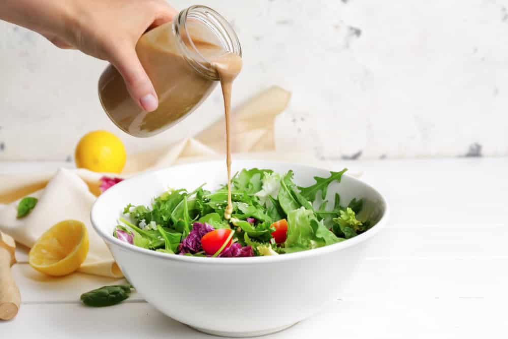 Woman pouring tasty tahini from jar onto vegetable salad in bowl