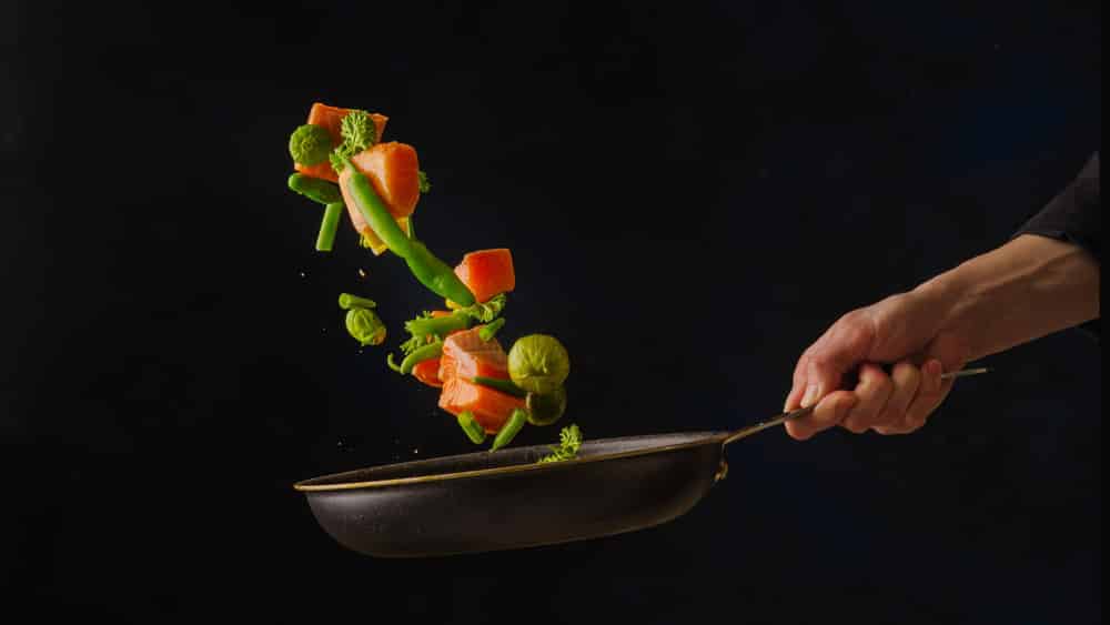 A frying pan in the chef's hand with pieces of red fish and vegetables