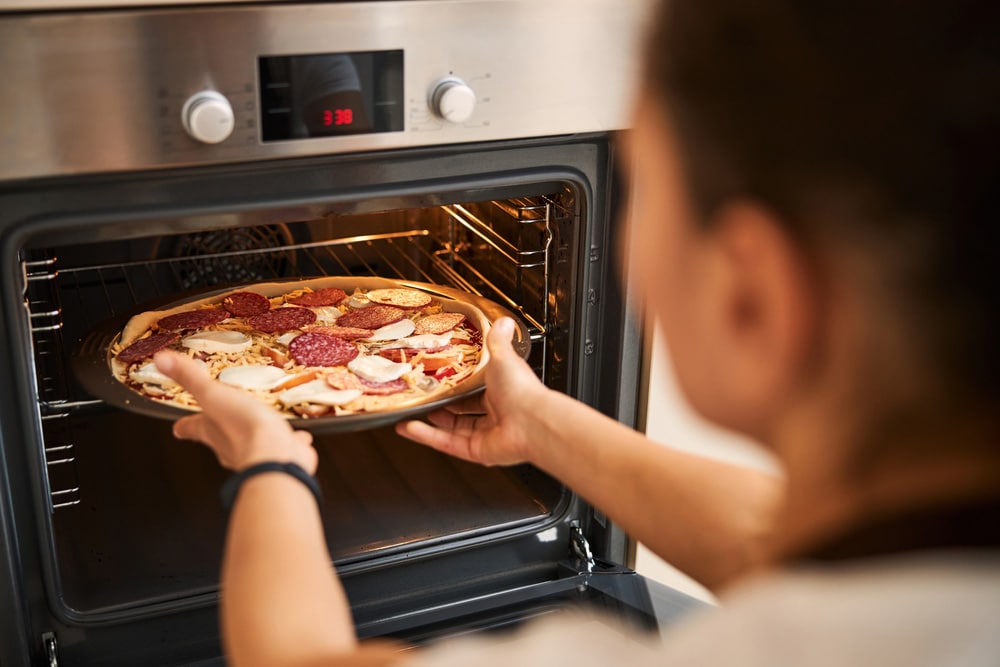 Unrecognized lady carefully putting a baking pan with pizza into the hot oven