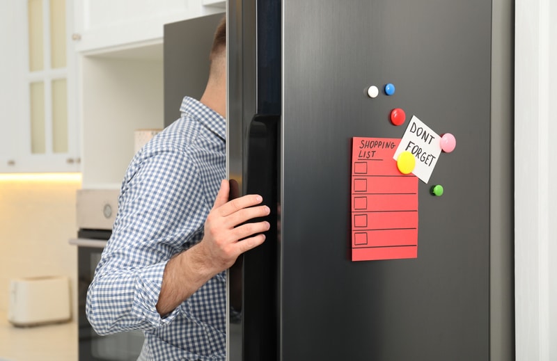 Man opening refrigerator door with shopping list, note and magnets in kitchen