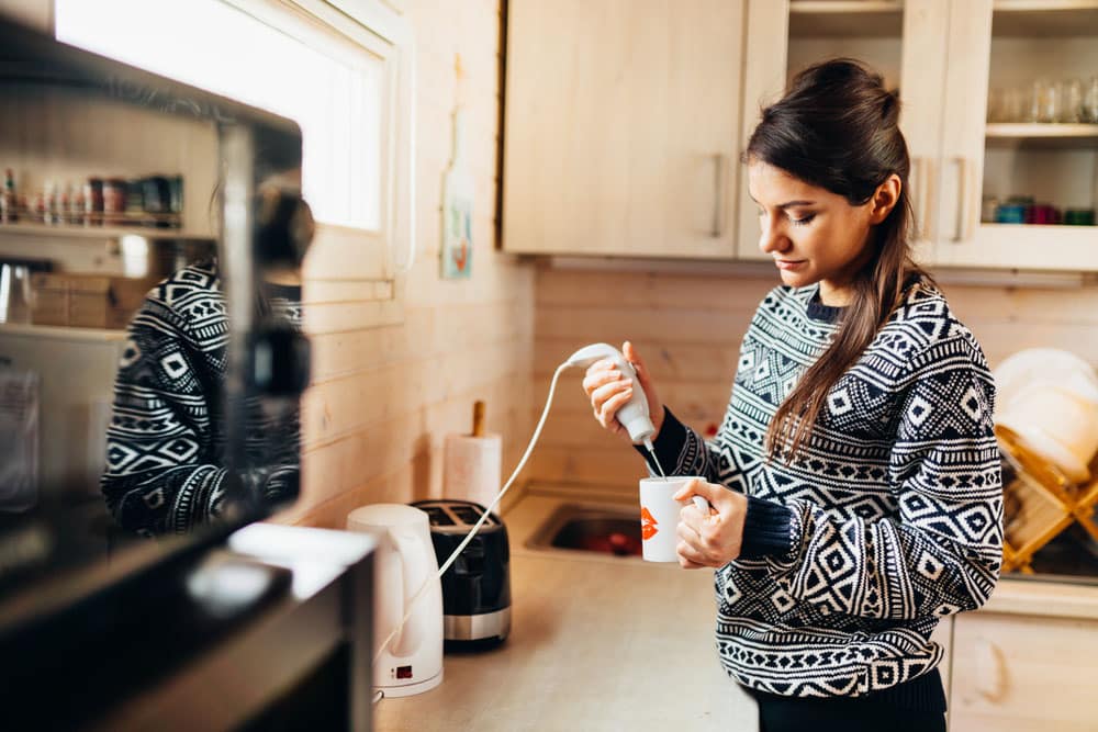 Woman making coffee drink at home kitchen using electric milk drink mixer whisk for frothy cream whipped coffee