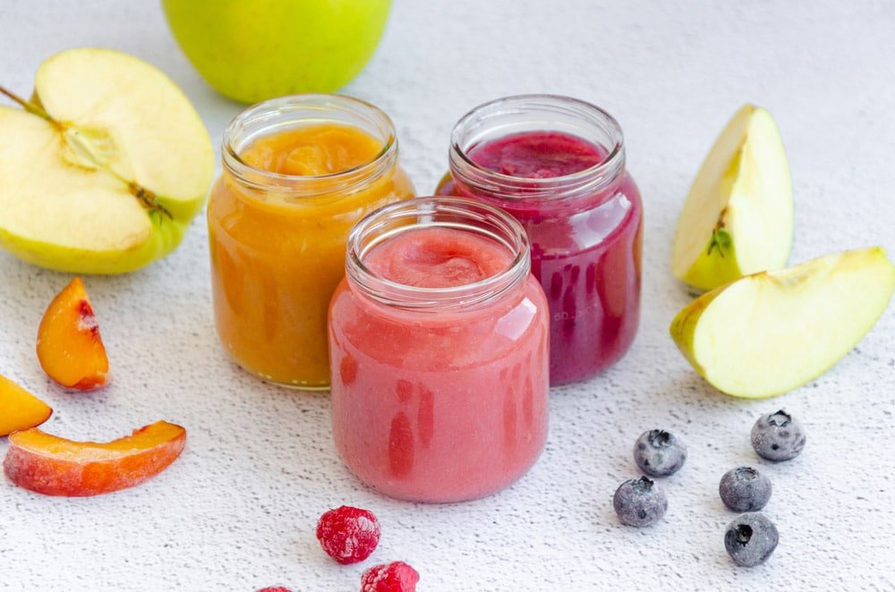 Variety of apple puree or applesauce with frozen peach, raspberries and blueberries in three glass jars on a light background