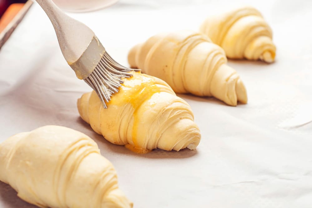 Baker brushing egg wash on uncooked croissants pastry roll