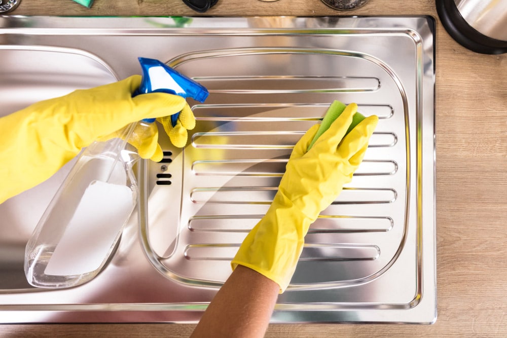 Person's Hand Cleaning Stainless Steel Sink With Sponge And Spray Bottle