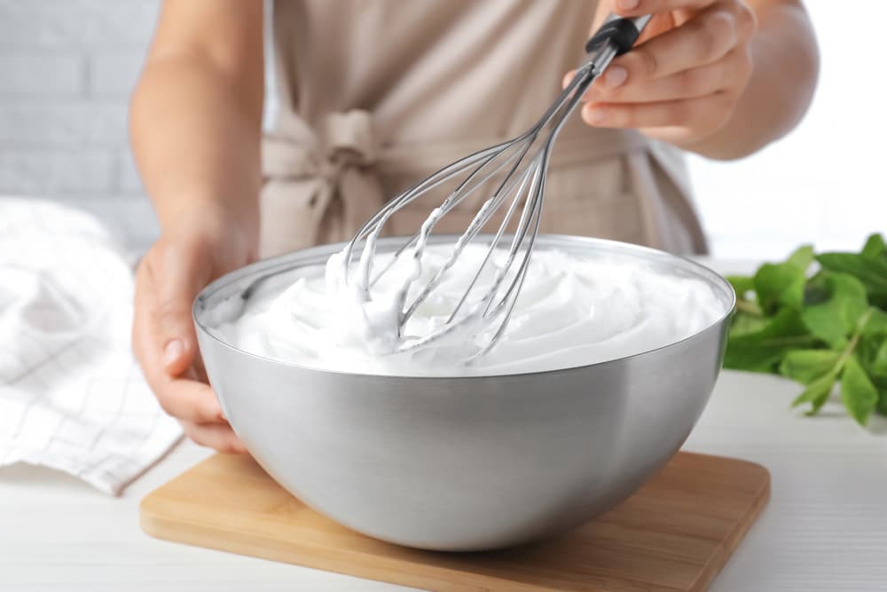 Woman whipping white cream with balloon whisk at wooden table