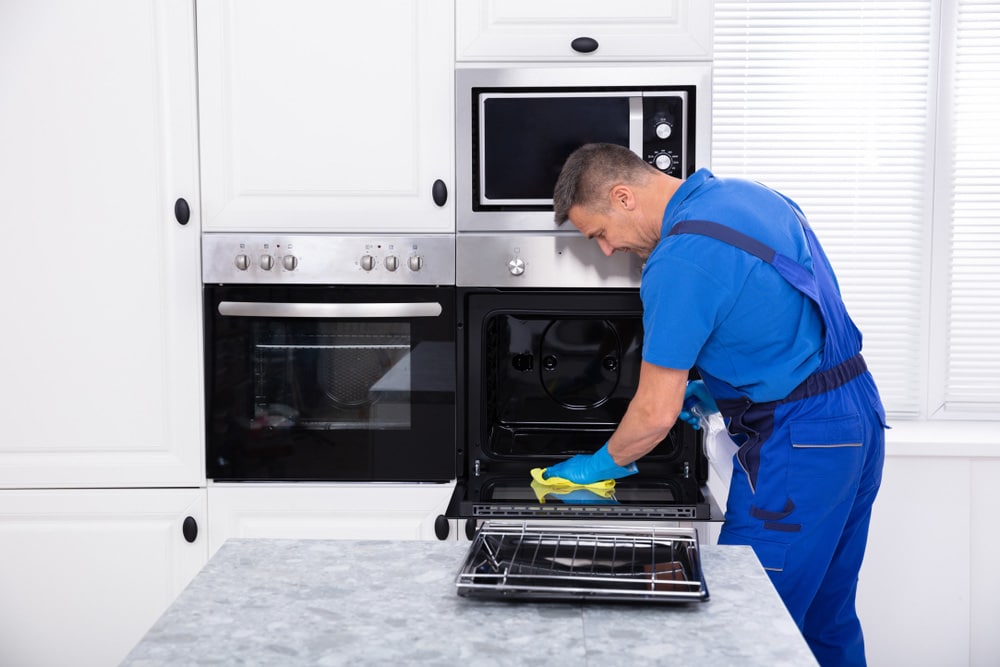Close-up Of A Smiling Male Janitor Cleaning Oven With Yellow Napkin In The Kitchen