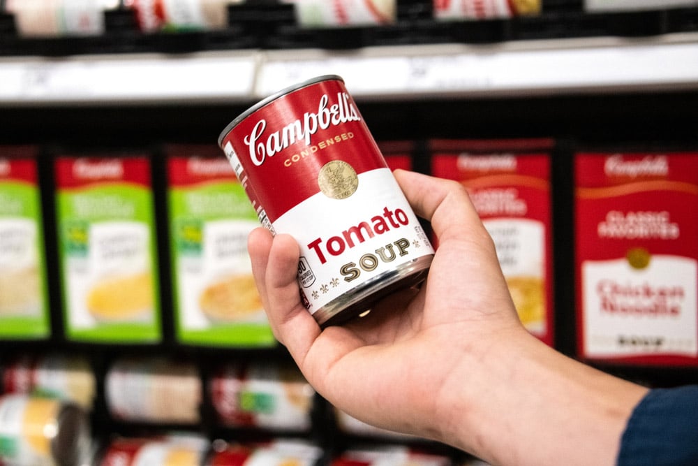 Shoppers hand holding a tin can of Campbells Brand Tomato soup