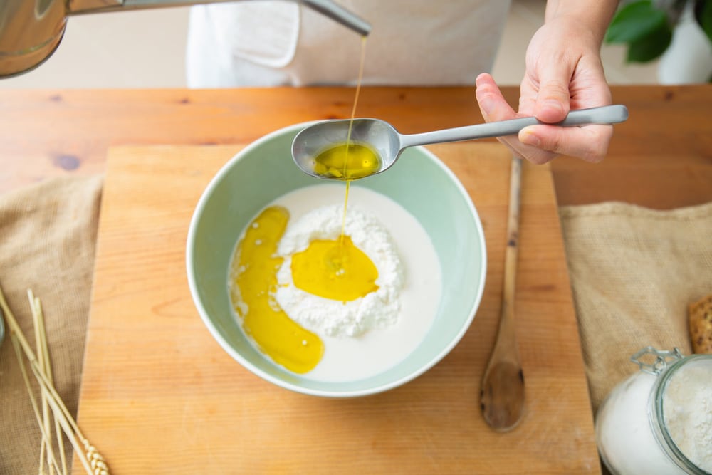 Skilled baker pouring olive oil from spoon to wheat flour in ceramic bowl
