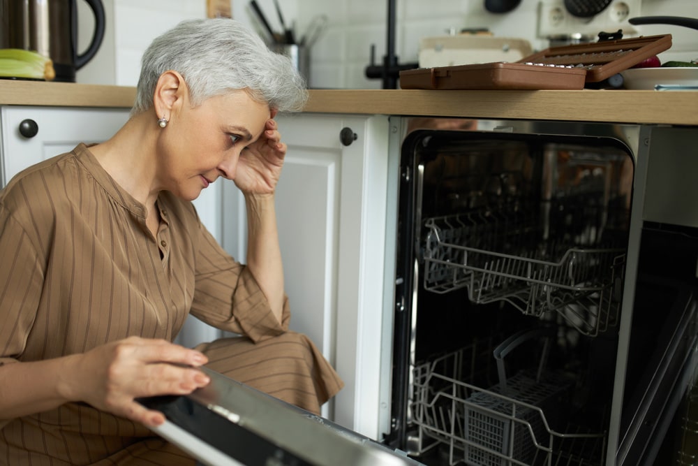Upset middle-aged housewife sitting at broken dishwasher