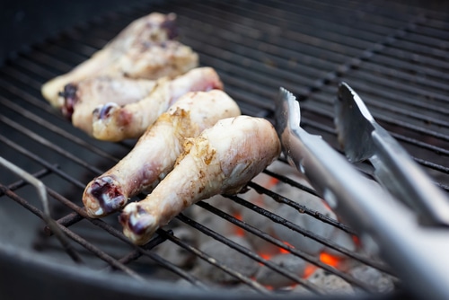 A closeup view of a charcoal barbecue with raw chicken drumsticks and tongs on the grill