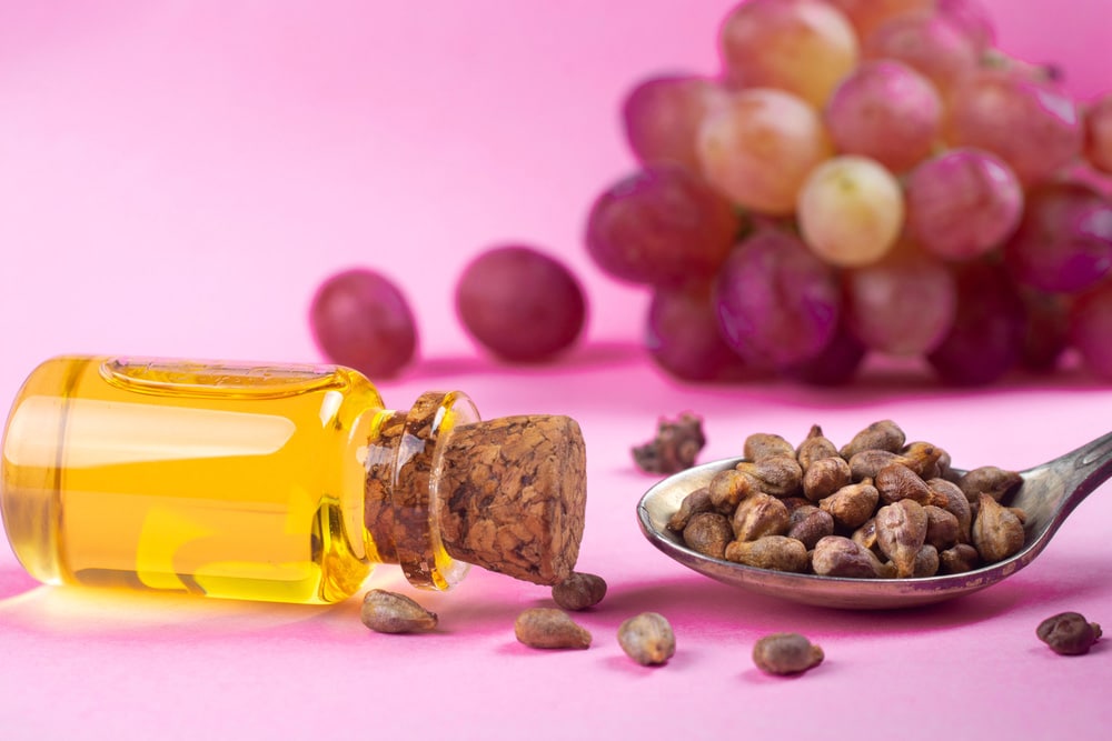 Grape seed oil in a glass jar and fresh grapes on a pink background