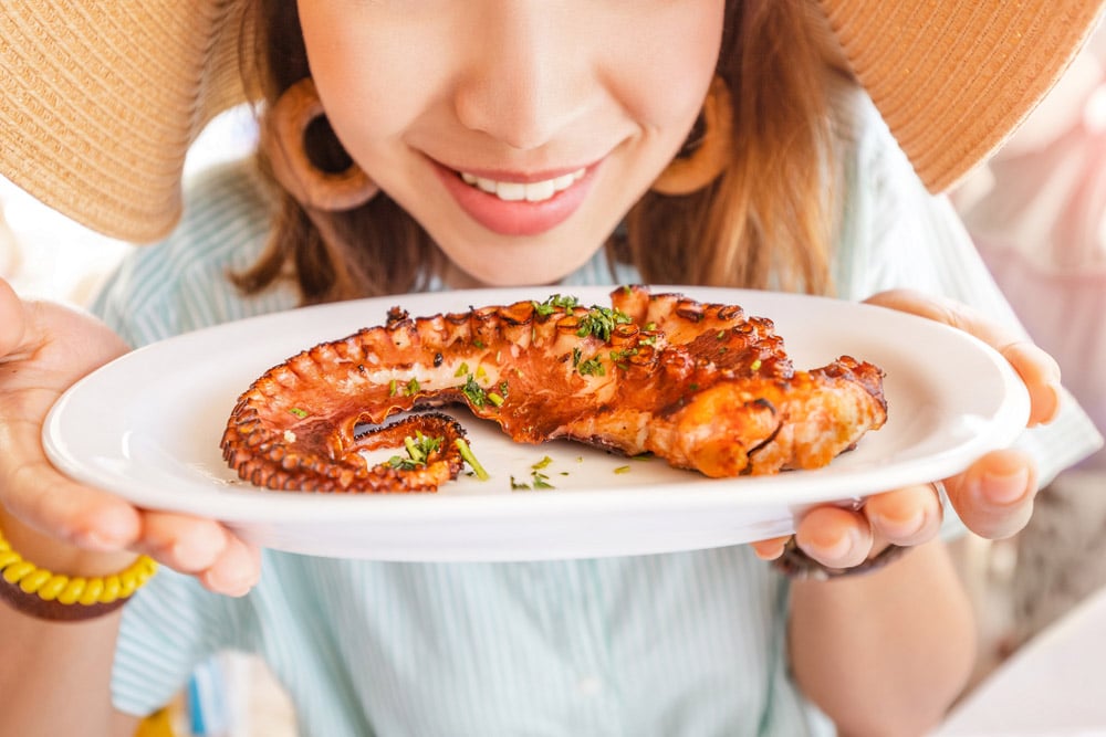 Happy asian woman in hat eating delicious grilled octopus in seafood restaurant
